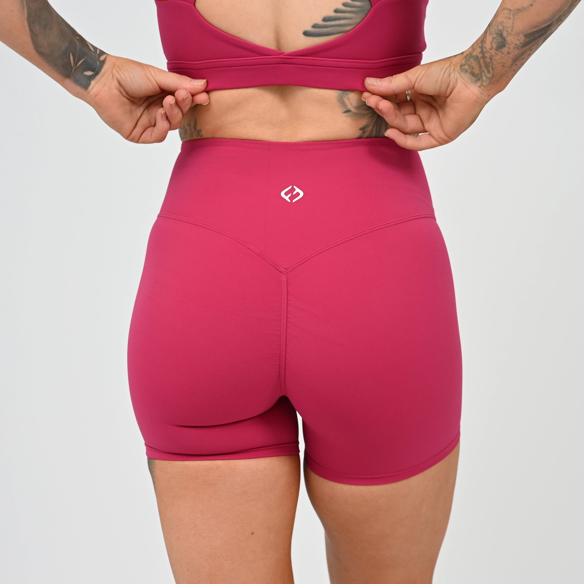 Rear view of a model wearing Hybryd shorts and a matching sports bra, with hands pulling the sports bra straps down. The shorts feature a small white Hybryd logo on the back waistband. Hidden Cinch bum bootys.