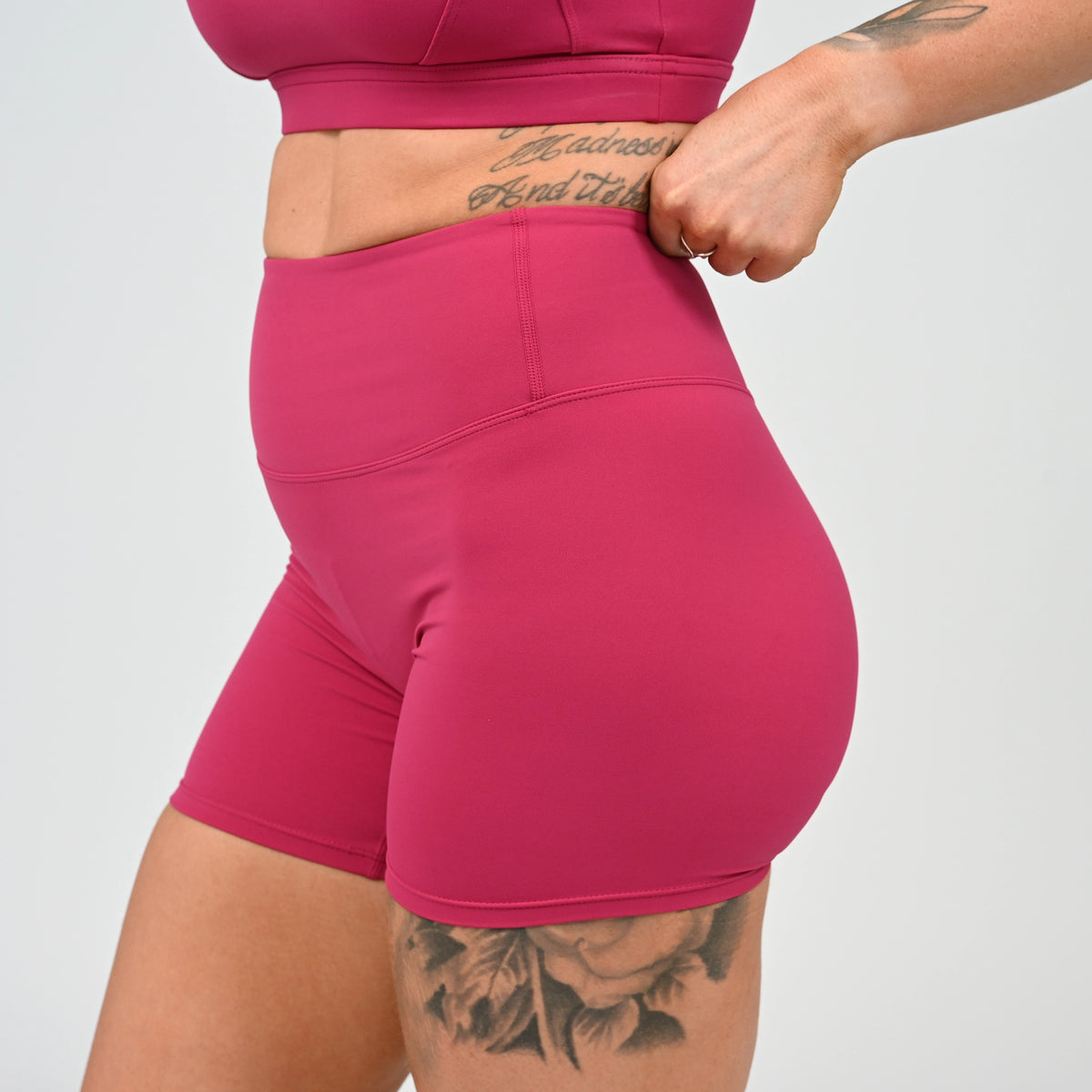 Side view of a Hyrox model wearing Hybryd shorts and a matching sports bra, with hands pulling up the booty shorts. The shorts feature a small white Hybryd logo on the back waistband and a Hidden Cinch bum bootys.