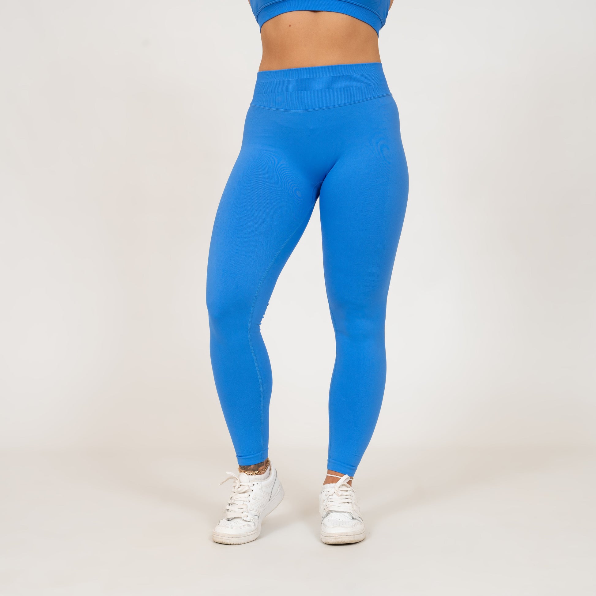 A pair of blue Hybryd leggings with a high-waisted fit and ruched stitching at the back, being worn with white sneakers.