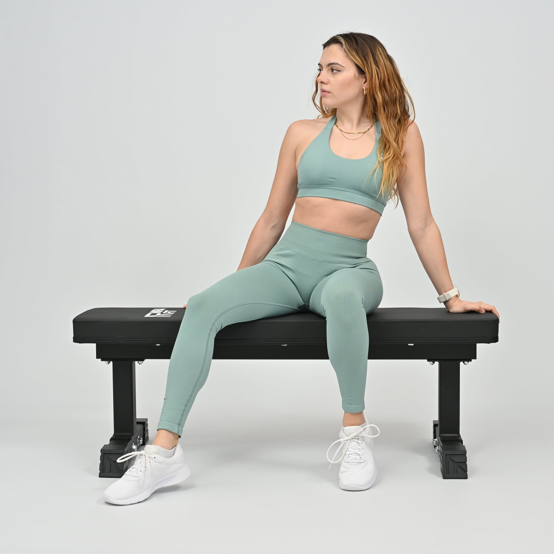 Woman in green athletic wear sitting on a black weight bench against a plain background