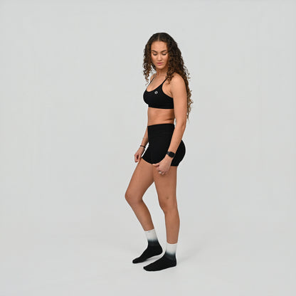 Woman wearing a black minimal Hybryd sports bra and booty shorts set with ombre sport socks on a light gray background