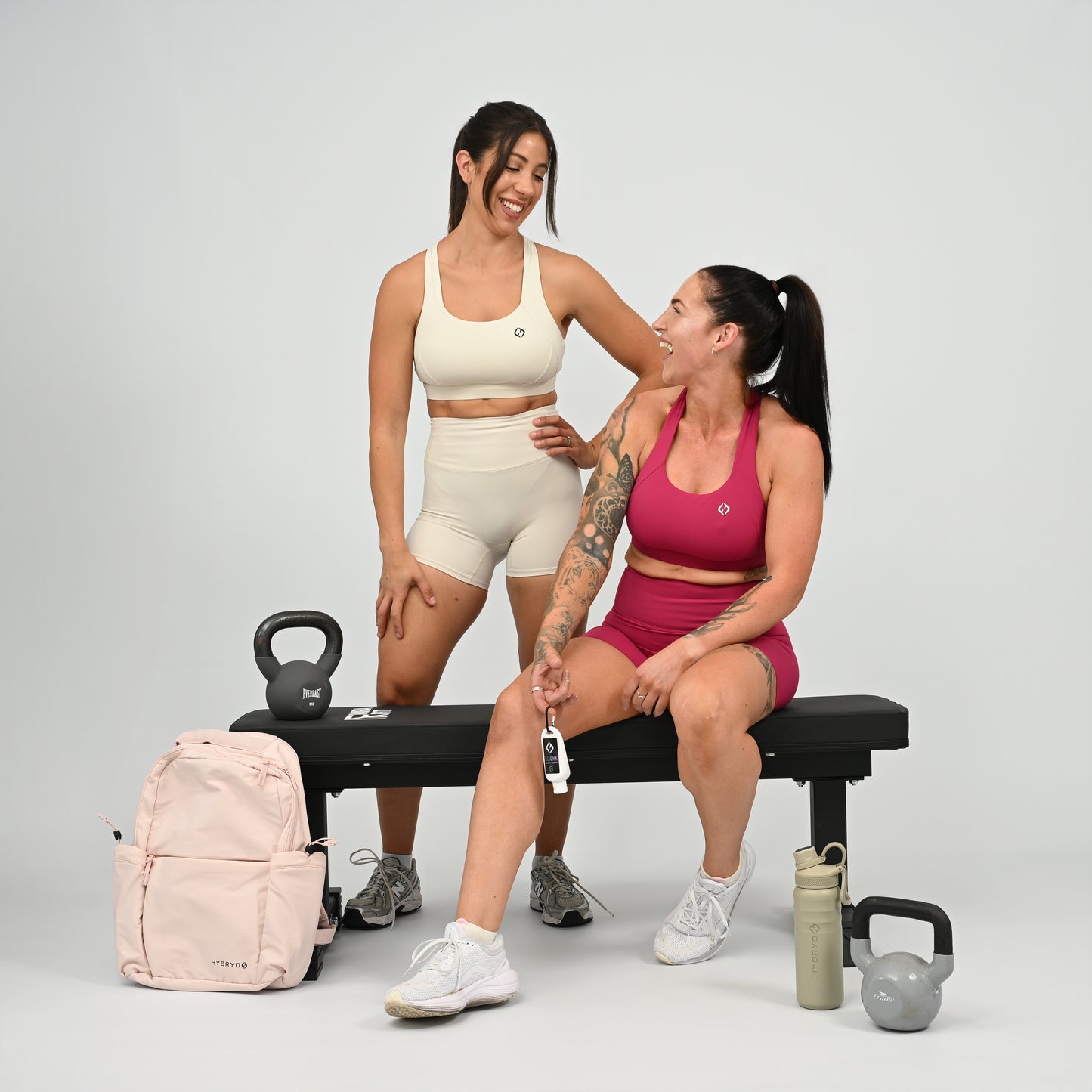 Two women in matching athletic wear pose on and beside a black weight bench in a studio setting. One wears a cream sports bra and shorts set, and the other wears a pink sports bra and shorts set. From gymwear brand Hybryd.