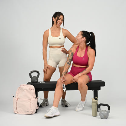 Two women in matching athletic wear pose on and beside a black weight bench in a studio setting. One wears a cream sports bra and shorts set, and the other wears a pink sports bra and shorts set. From gymwear brand Hybryd.