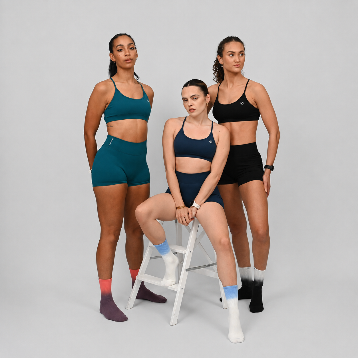 Three women model athletic wear in a studio setting. One sits on a white stool in the center wearing a navy sports bra and shorts, while the other two stand behind her in teal and black sets.