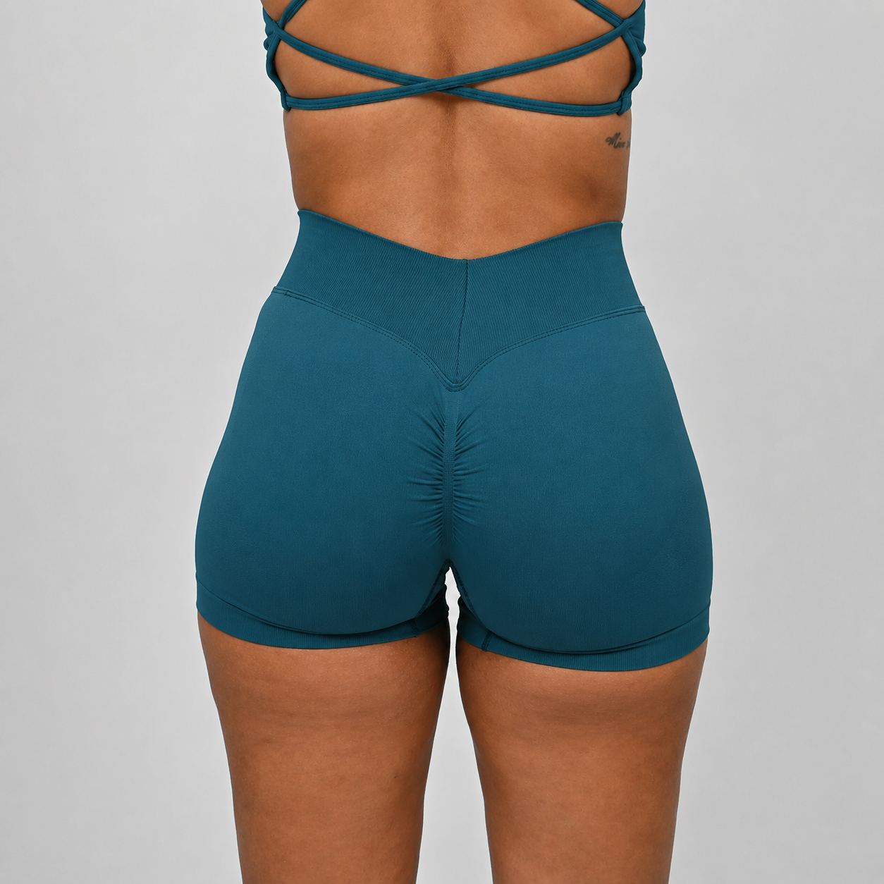 Close-up back view of a model wearing high-waisted teal hybryd booty shorts with a wide ribbed waistband and matching teal sports bra straps visible at the top, against a light gray studio background.