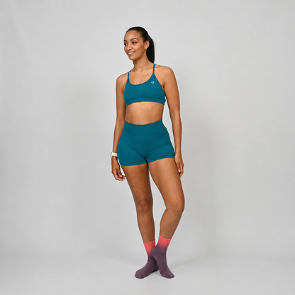 A woman modeling a teal matching set consisting of a sports bra and matching high-waisted shorts, paired with pink and purple socks, against a light gray studio background. rom activewear brand Hybryd.