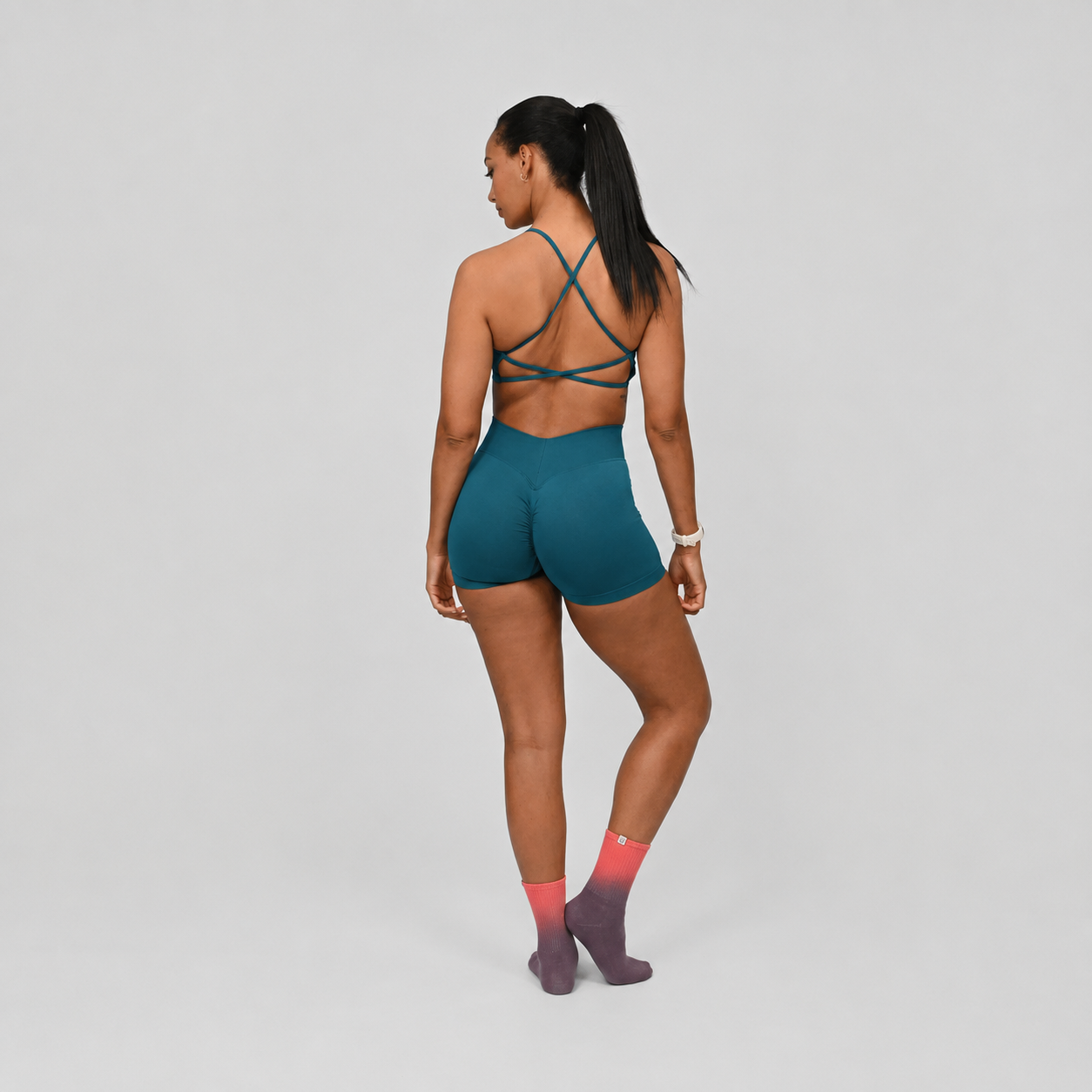 A woman is shown from behind wearing a teal athletic sports bra with crisscross straps and matching teal high-waisted shorts, paired with pink and purple socks, against a light gray studio background. From gymwear brand Hybryd.
