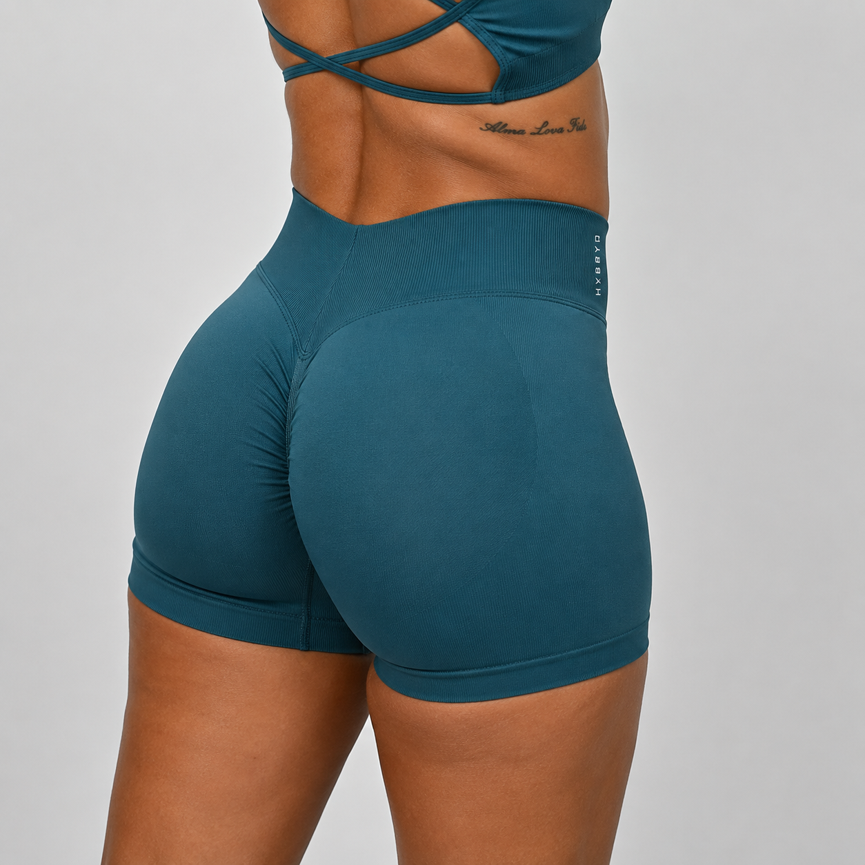 Close-up back view of a model wearing high-waisted teal hybryd booty shorts with a wide ribbed waistband and matching teal sports bra straps visible at the top, against a light gray studio background.