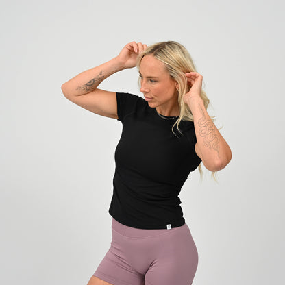 Woman wearing a black stretchy, fitted  gym  t-shirt with a Hybryd logo and booty shorts against a plain background