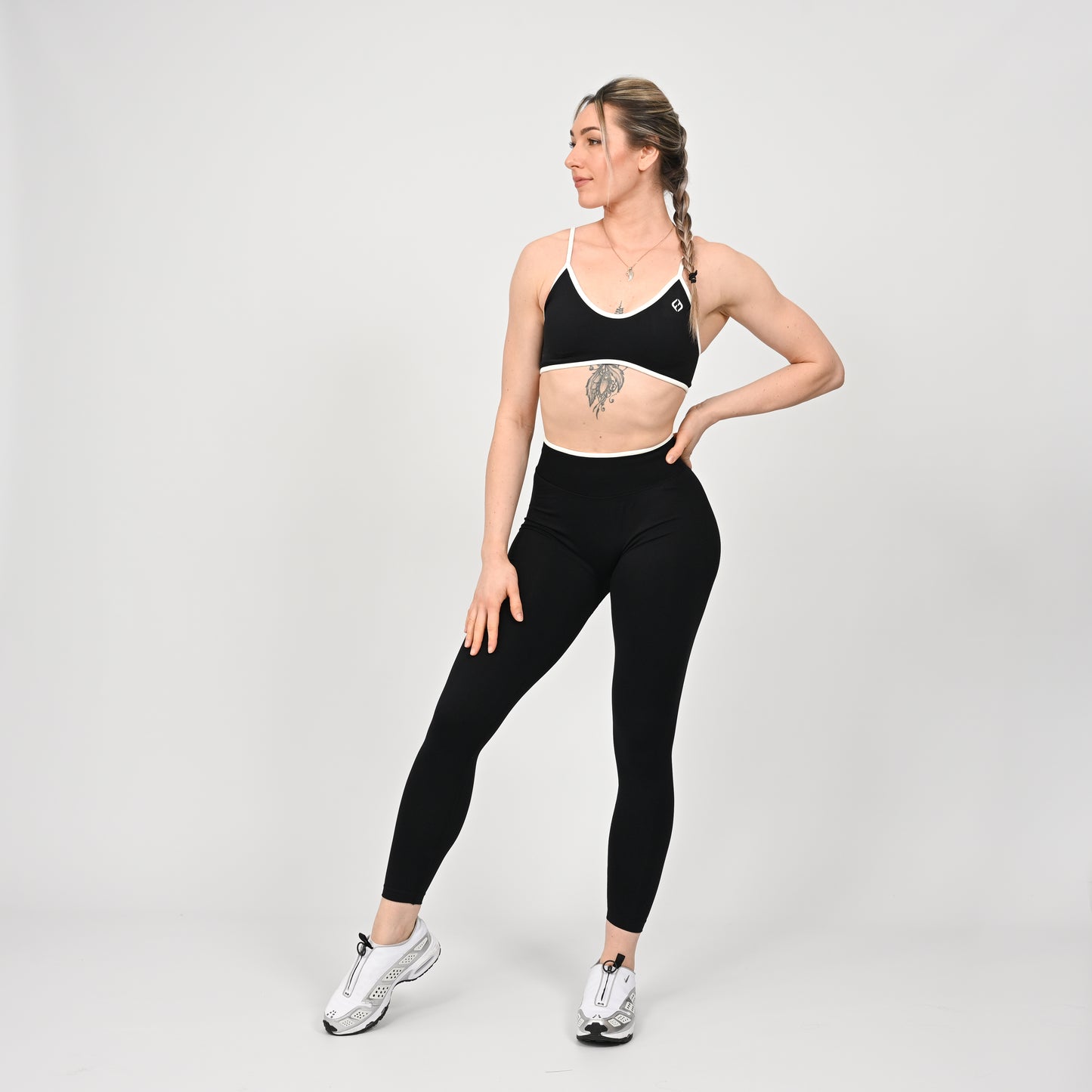 Crossfit girl wearing a black minimal sports bra with white piping and removable pads. Worn with matching cinch bum leggings. Front view on a off white background.