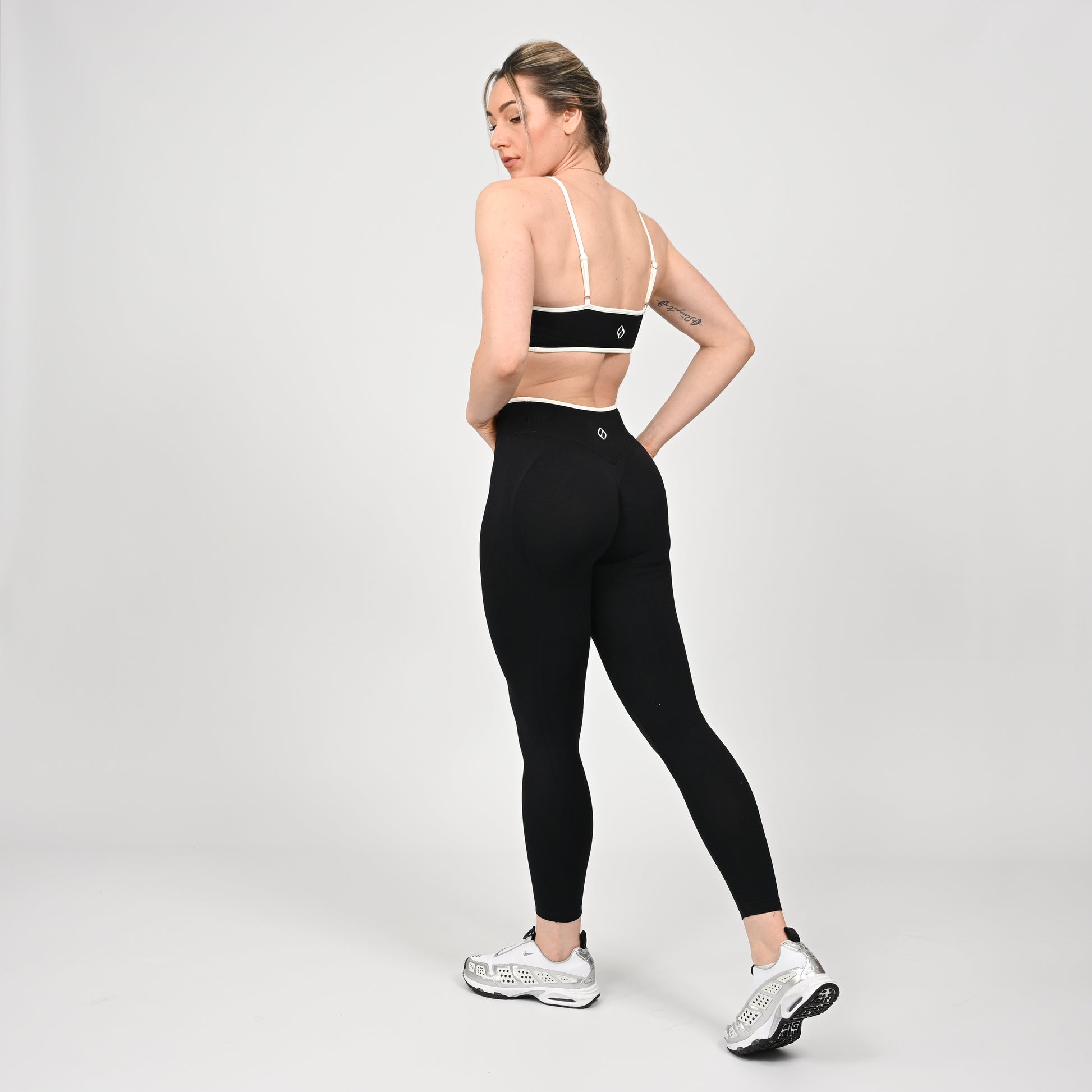 Crossfit girl wearing a black minimal sports bra with white piping and removable pads. Worn with matching cinch bum leggings. Rear view on a off white background.