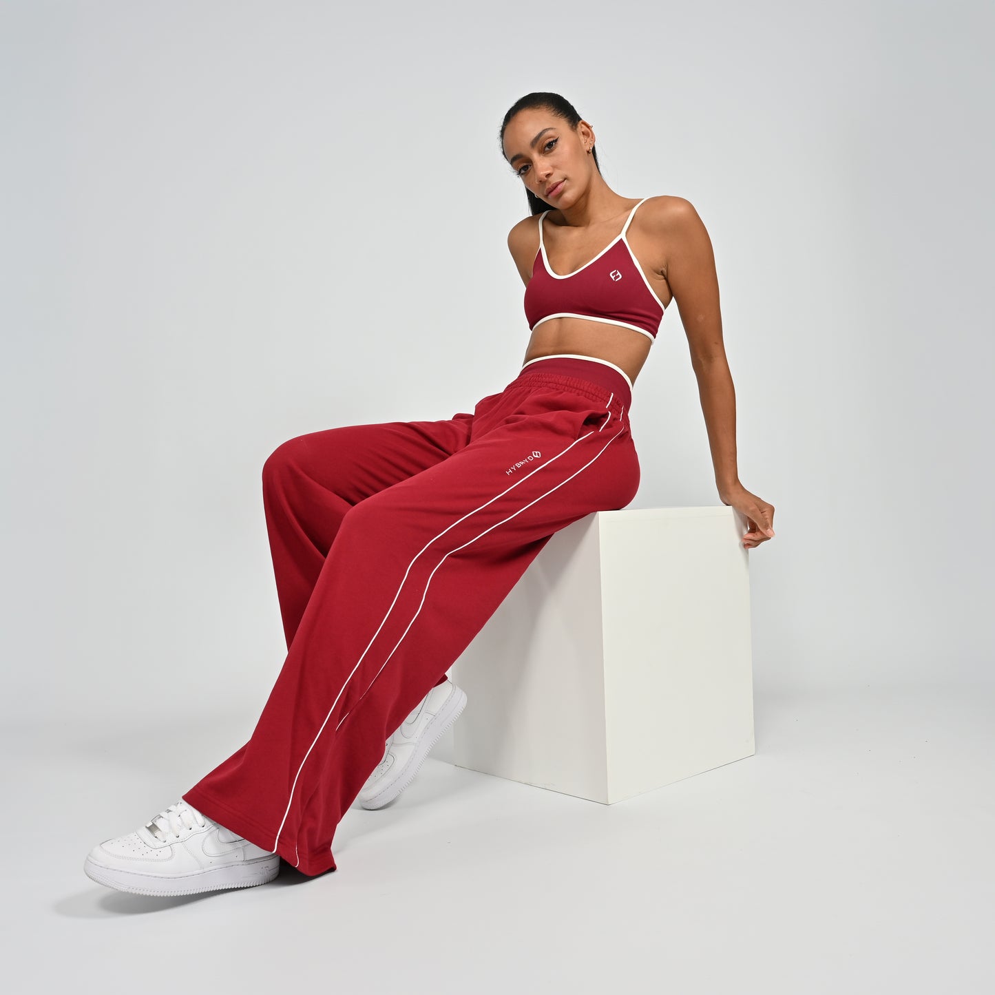 Gym Athlete wearing a crimson red Hybryd minimal sports bra and wide leg joggers with white piping on a light gray background