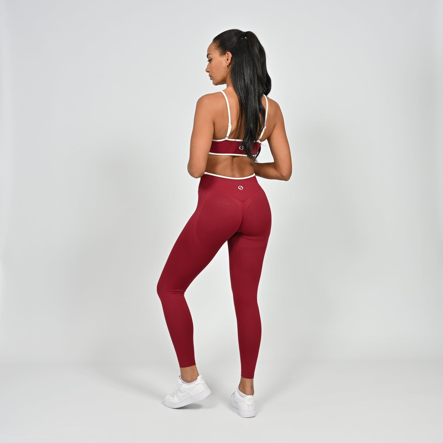 Gym Athlete wearing a crimson red Hybryd minimal sports bra and matching cinch bum leggings with white piping on a light gray background rear view