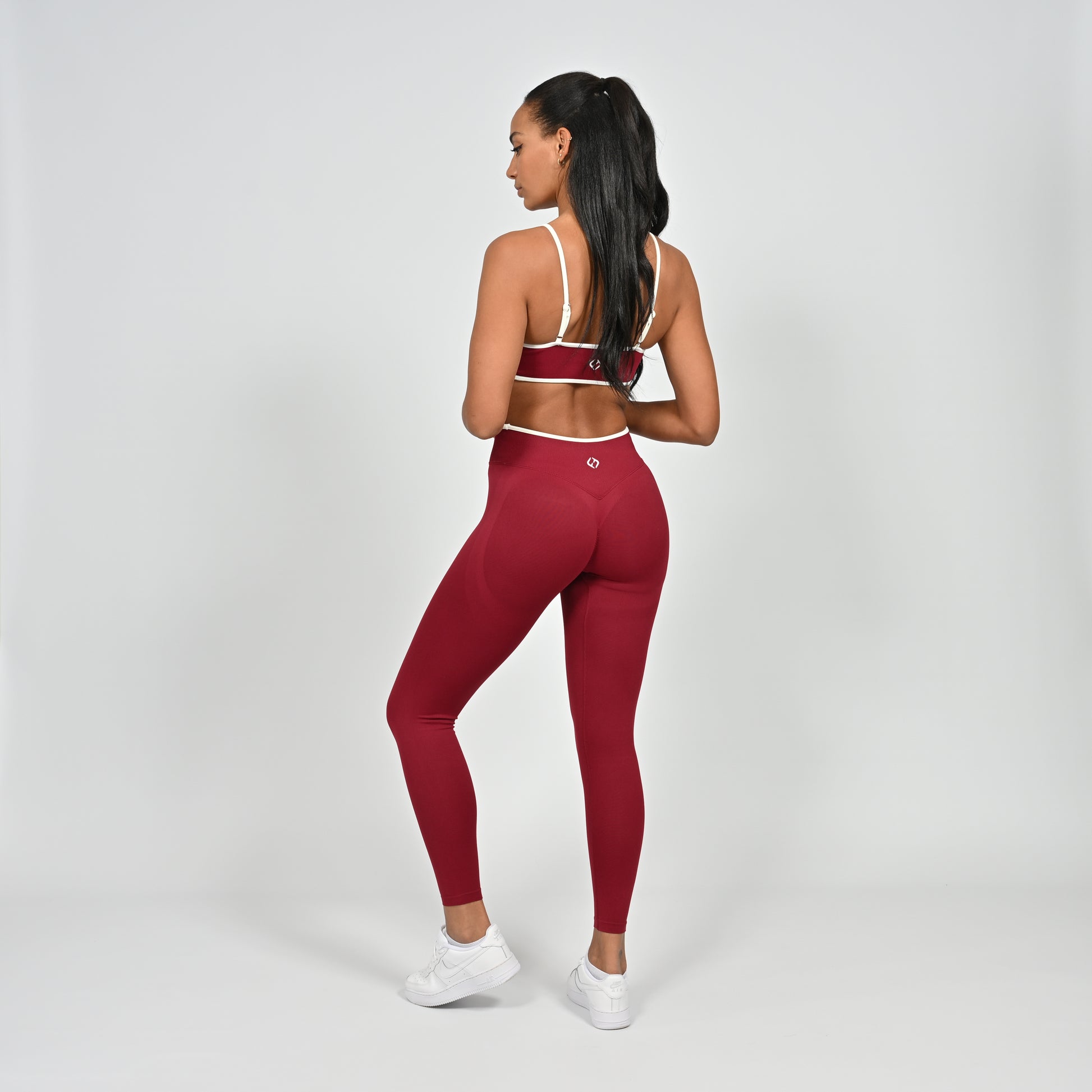 Gym Athlete wearing a crimson red Hybryd minimal sports bra and matching cinch bum leggings with white piping on a light gray background rear view
