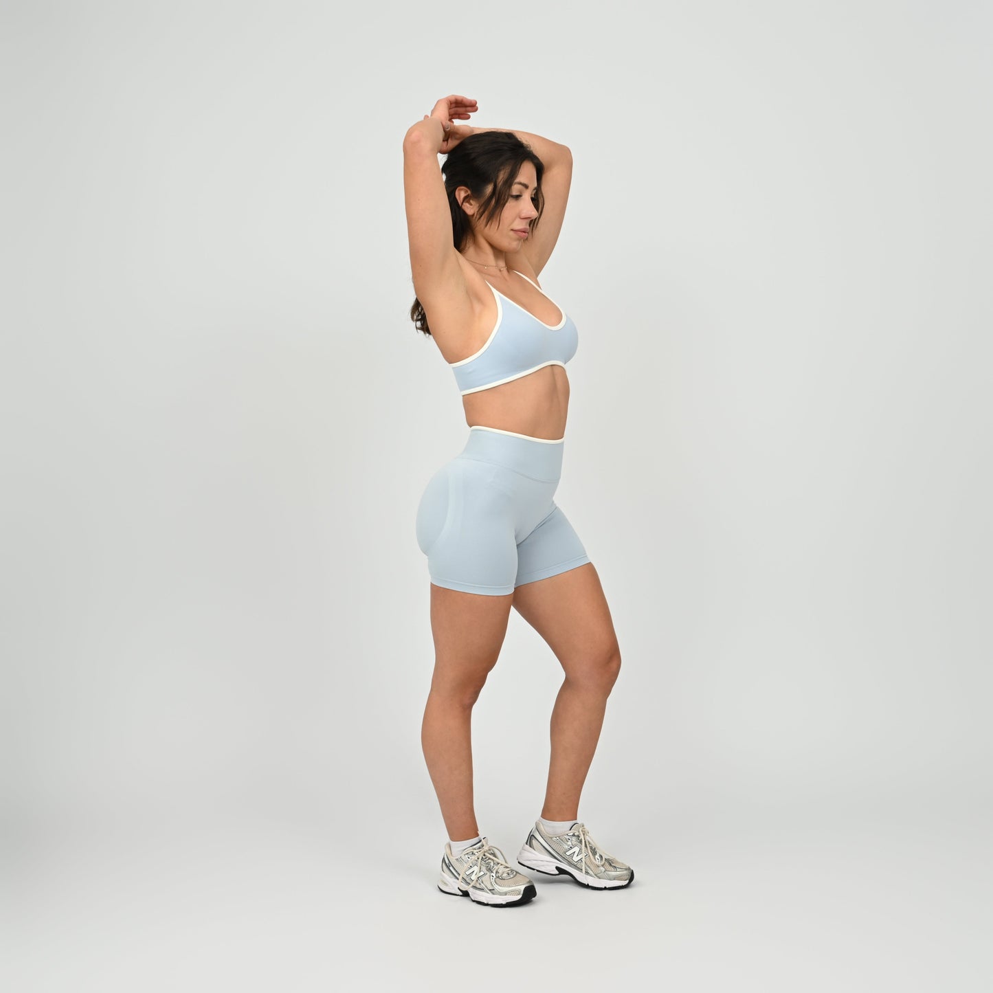 Crossfit girl wearing a powder blue minimal sports bra with white piping and removable pads. Worn with matching cinch bum bootie shorts. Side view on a white background. Made by the gymwear brand Hybryd.