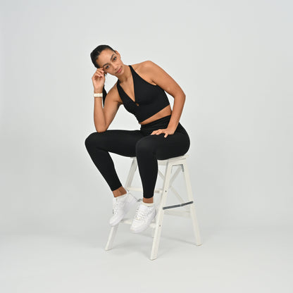 Woman in black Hybryd athletic wear sitting on a white stool against a light gray background