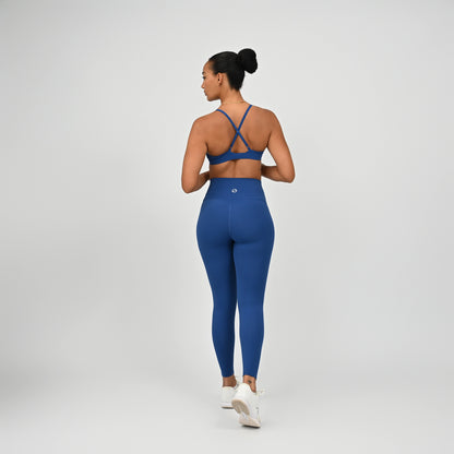 A woman is shown from behind wearing a matching Hybryd royal blue activewear set consisting of a sports bra with criss-cross straps and high-waisted leggings, paired with white sneakers, against a light gray studio background.