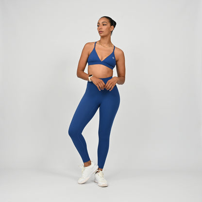 A woman modeling a matching sportswear set consisting of a royal blue sports bra with a Hybryd logo and high-waisted leggings, paired with white sneakers, against a light gray studio background.