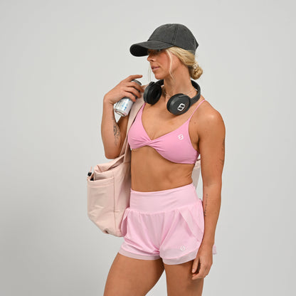 Woman in a pink sportswear matching set with a grey Hybryd cap and headphones, holding a water bottle and a bag on a light gray background.