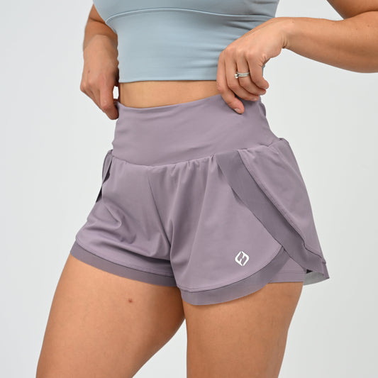 A close-up of a model wearing high-waisted running shorts in a dusty lavender-purple color. The shorts feature a wide waistband, side pockets, and a small white Hybryd logo on the lower left leg.