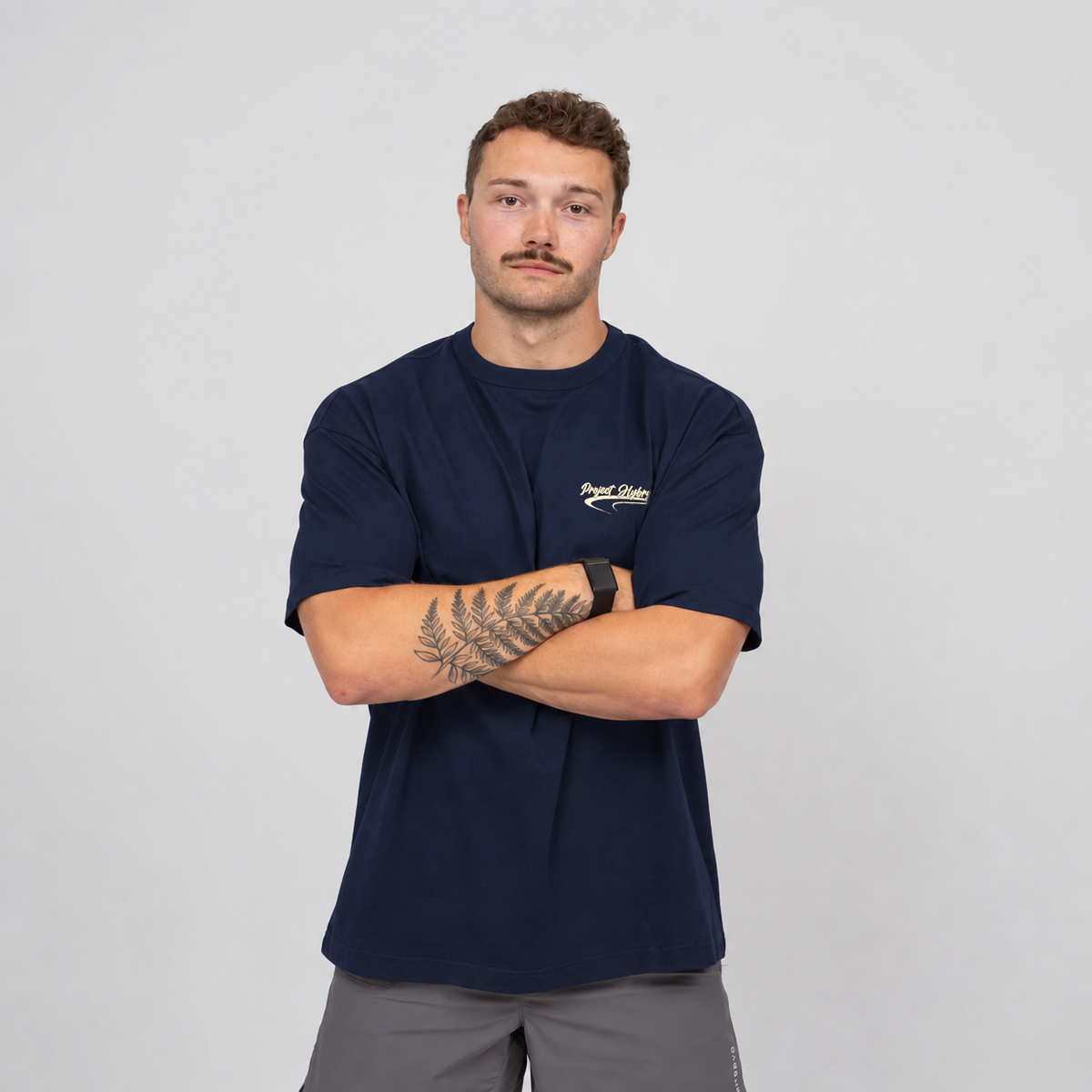 A man wearing a navy blue Hybryd t-shirt with a small white script logo on the left chest, paired with gray gym shorts, standing against a light gray background with his arms crossed.