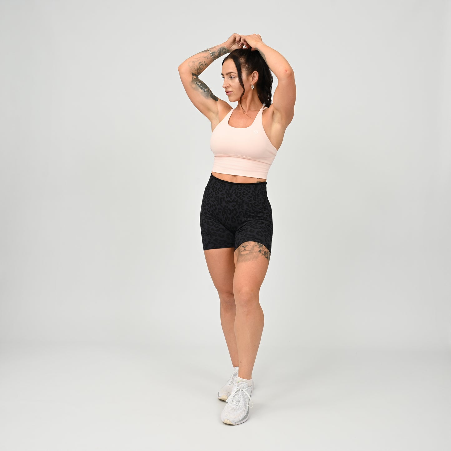 A woman modeling a light pink supportive gymwear Hybryd sports bra and black patterned athletic shorts against a light gray studio background.