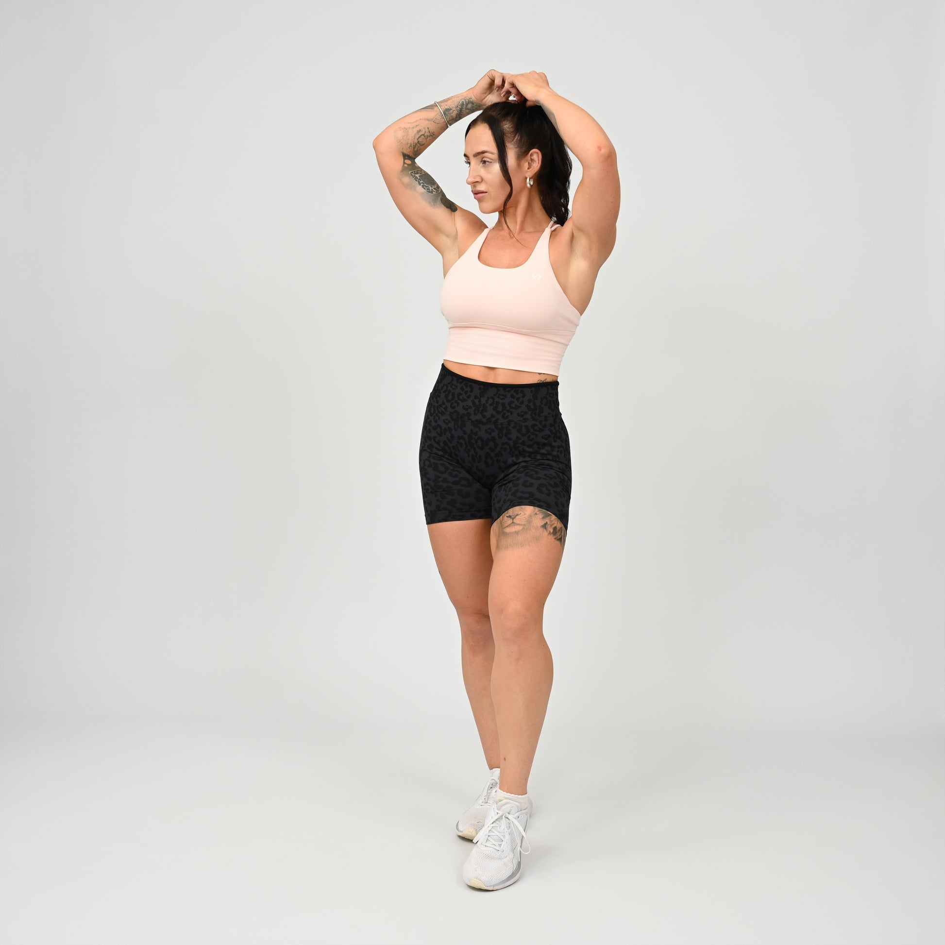 A woman modeling a light pink supportive gymwear Hybryd sports bra and black patterned athletic shorts against a light gray studio background.