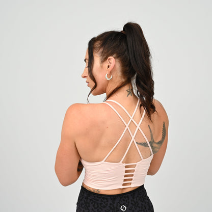 A athlete shown from the back wearing a pale blush pink Hybryd sports bra with crisscrossing straps and a cutout back design, paired with black patterned leggings against a light gray studio background.