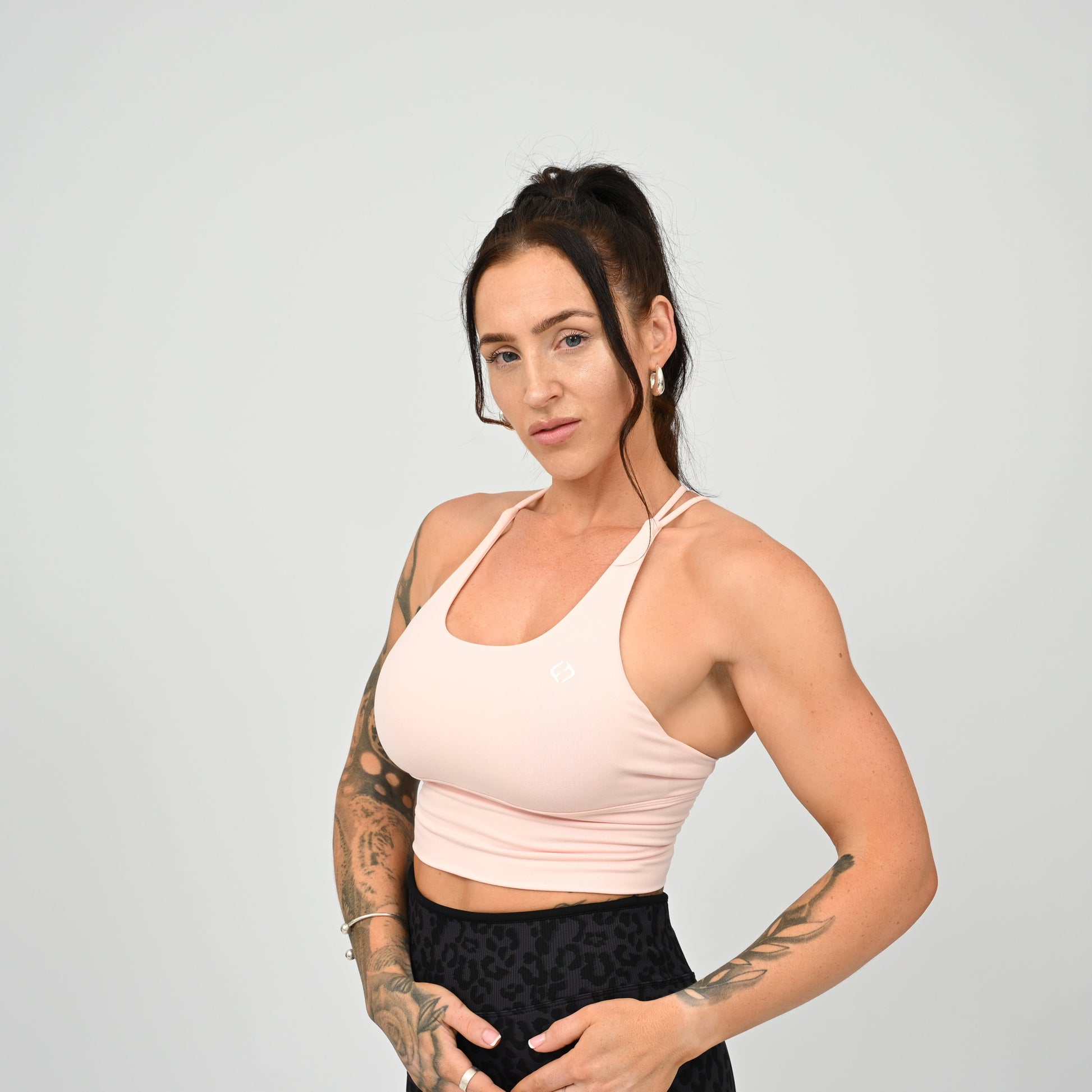 A CrossFit  athlete with tattoos modeling a light pink Hybryd sports bra and black patterned leggings against a solid light gray background.