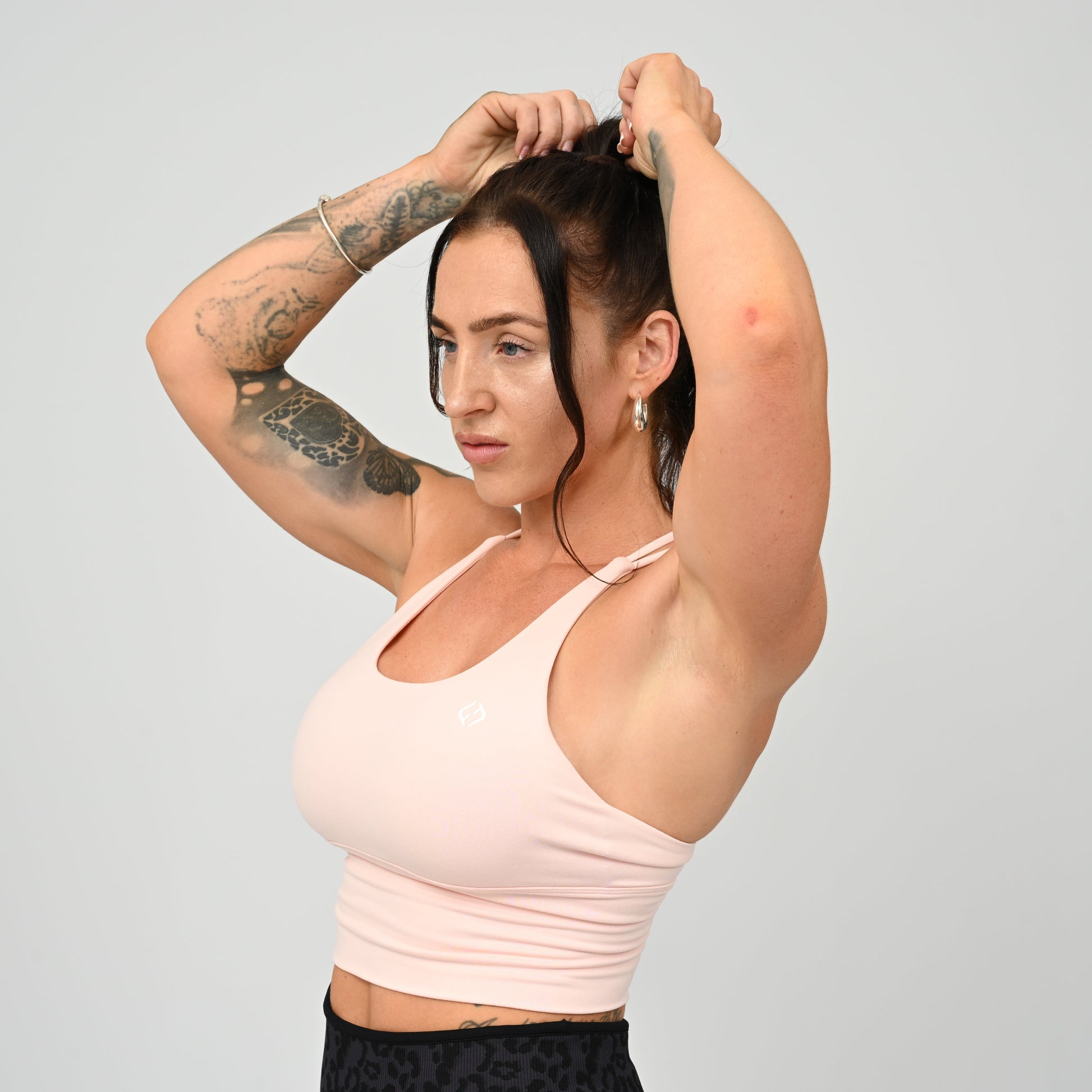 A crossfit athlete with tattoos adjusting her hair, wearing a pale pink Hybryd sports bra and black patterned leggings, set against a solid light gray background.