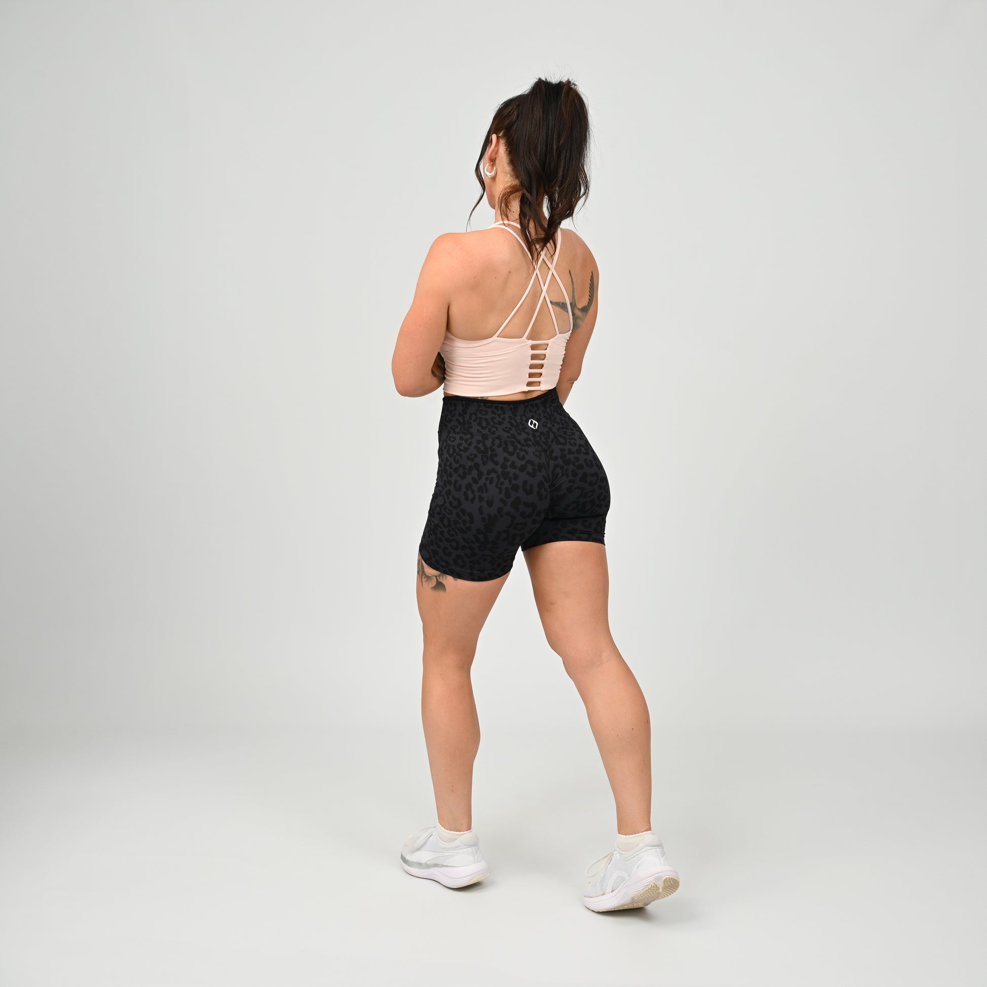 A woman is shown from behind wearing a light pink Hybryd athletic sports bra with crisscross straps and black patterned high-waisted gym shorts, paired with white sneakers, against a light gray studio background.