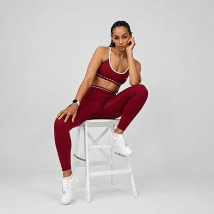 Hybryd yoga athlete in maroon athletic wear with piping with minimal bra sitting on a white stool against a gray background