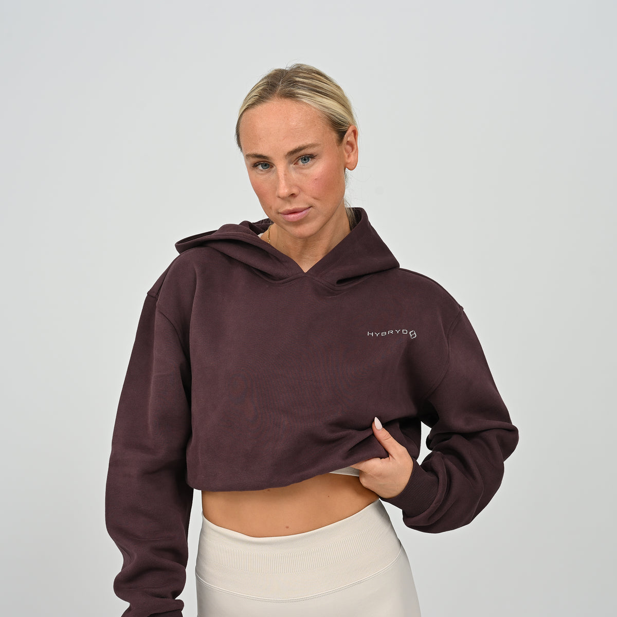 Front close-up view of model adjusting the hem of the aubergine Hybryd Barstow cropped hoodie