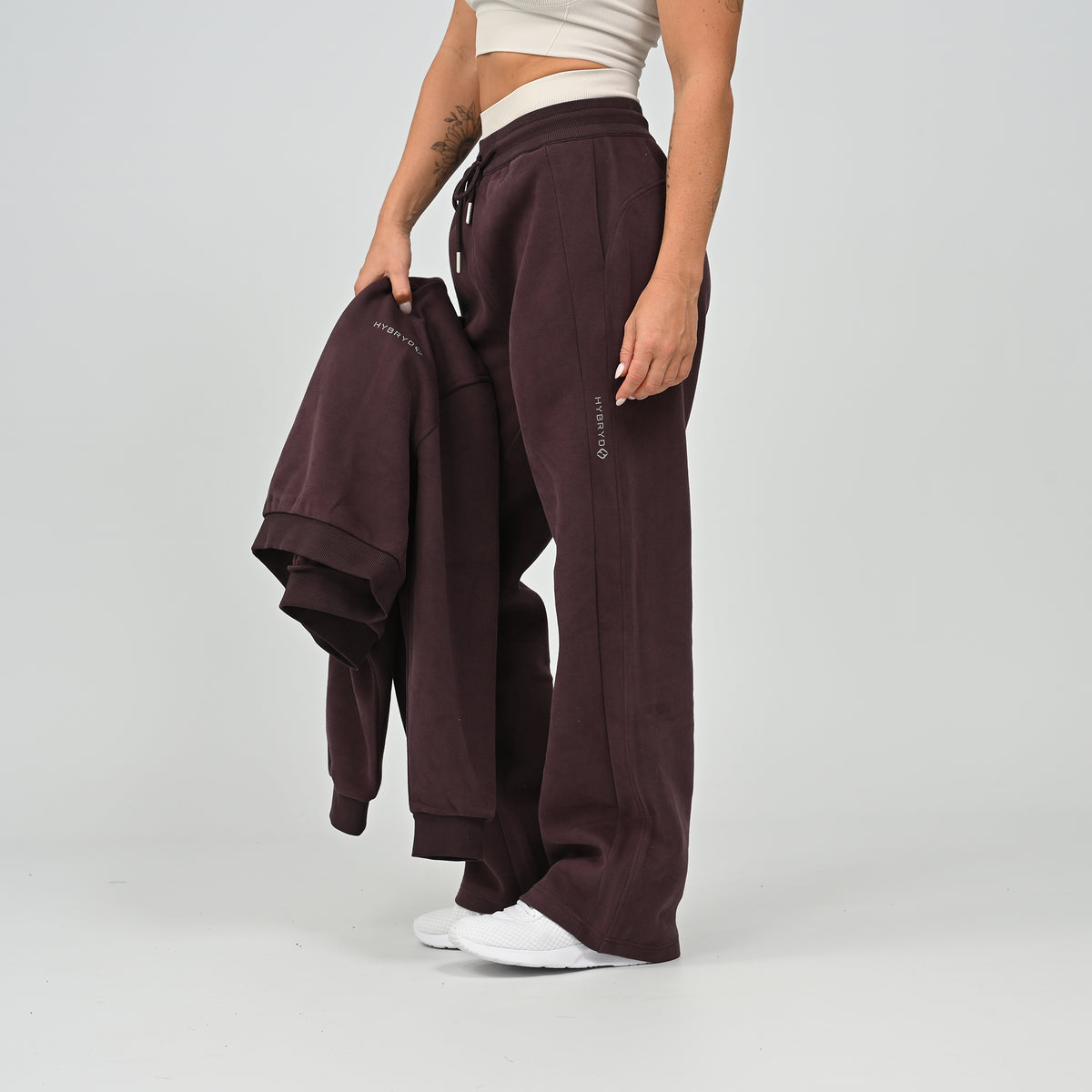 Model standing front view wearing the aubergine Hybryd Barstow wide leg joggers with cropped hoodie in hand