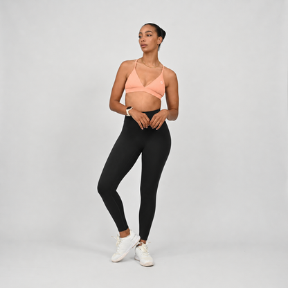 A crossfit athlete modeling a peach-coloured crossover sports bra and black high-waisted leggings with white sneakers, standing against a light gray studio background.