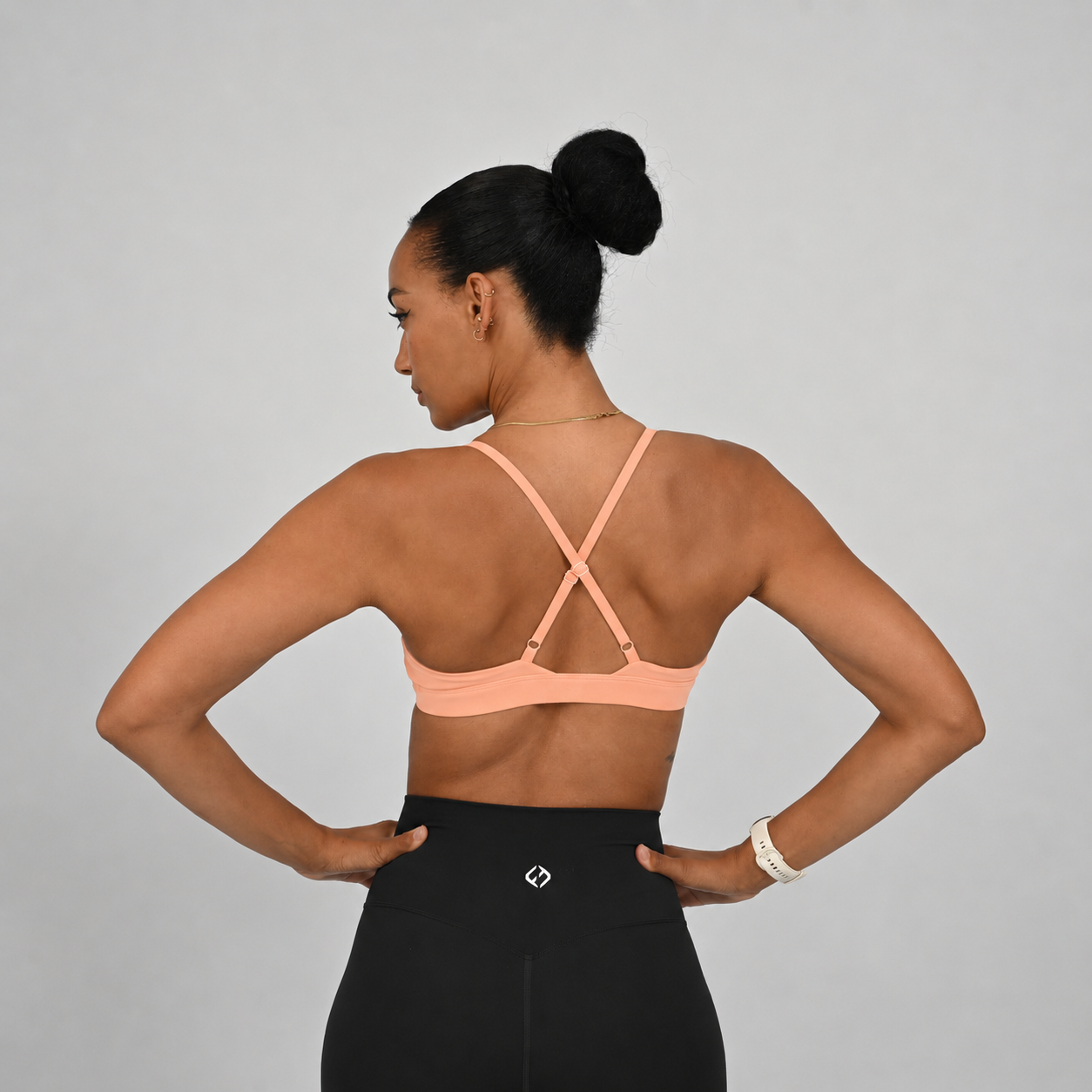 A woman shown from the back wearing a peach-colored sports bra with crisscross straps and black high-waisted leggings with a Hybryd logo, posing with hands on hips against a light gray studio background.
