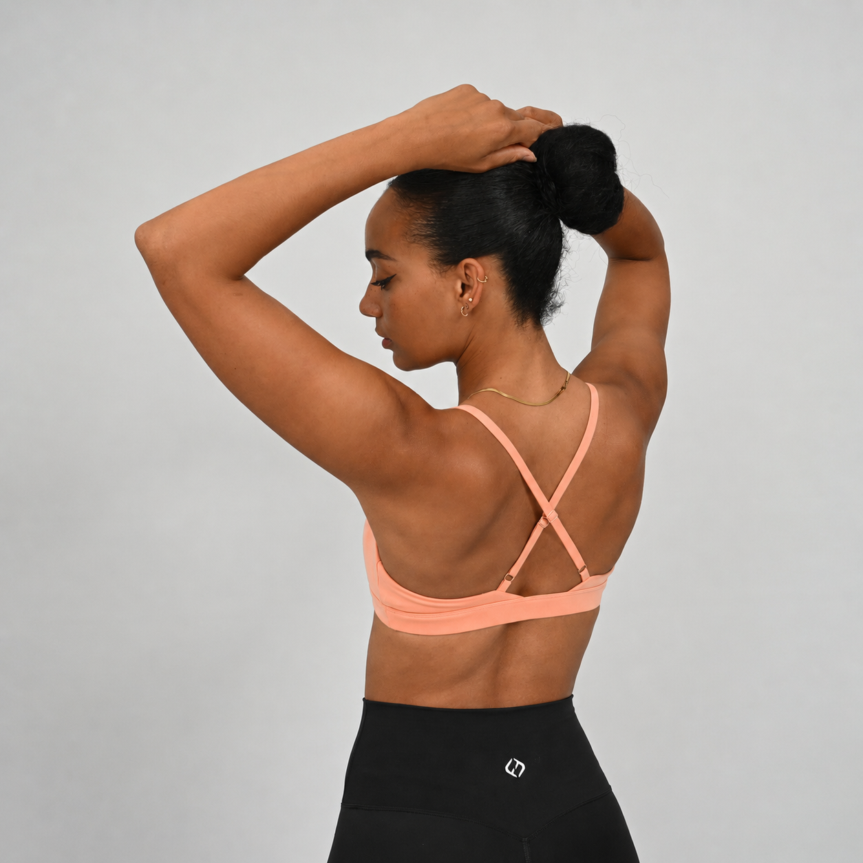 A woman wearing a peach-colored sports bra with crisscross adjustable back straps, shown from the back with Hybryd black leggings against a light gray studio background.