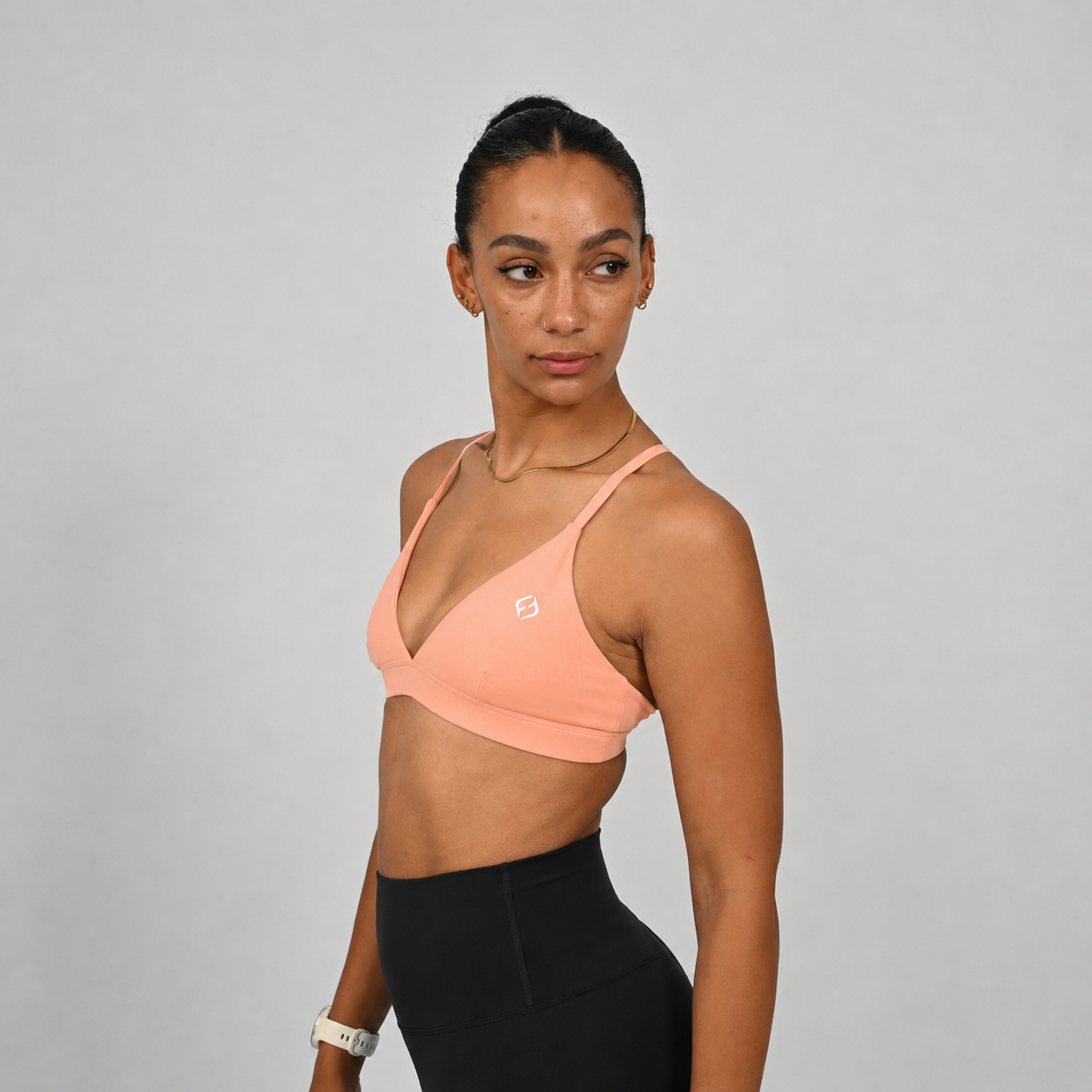 A woman wearing a peach-colored minimal Hybryd sports bra and black athletic leggings, photographed against a plain light gray studio background.