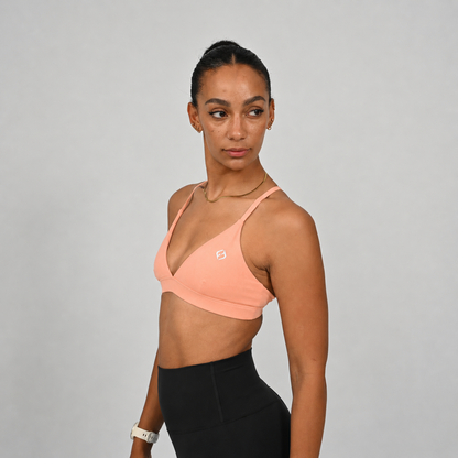 A woman wearing a peach-colored minimal Hybryd sports bra and black athletic leggings, photographed against a plain light gray studio background.