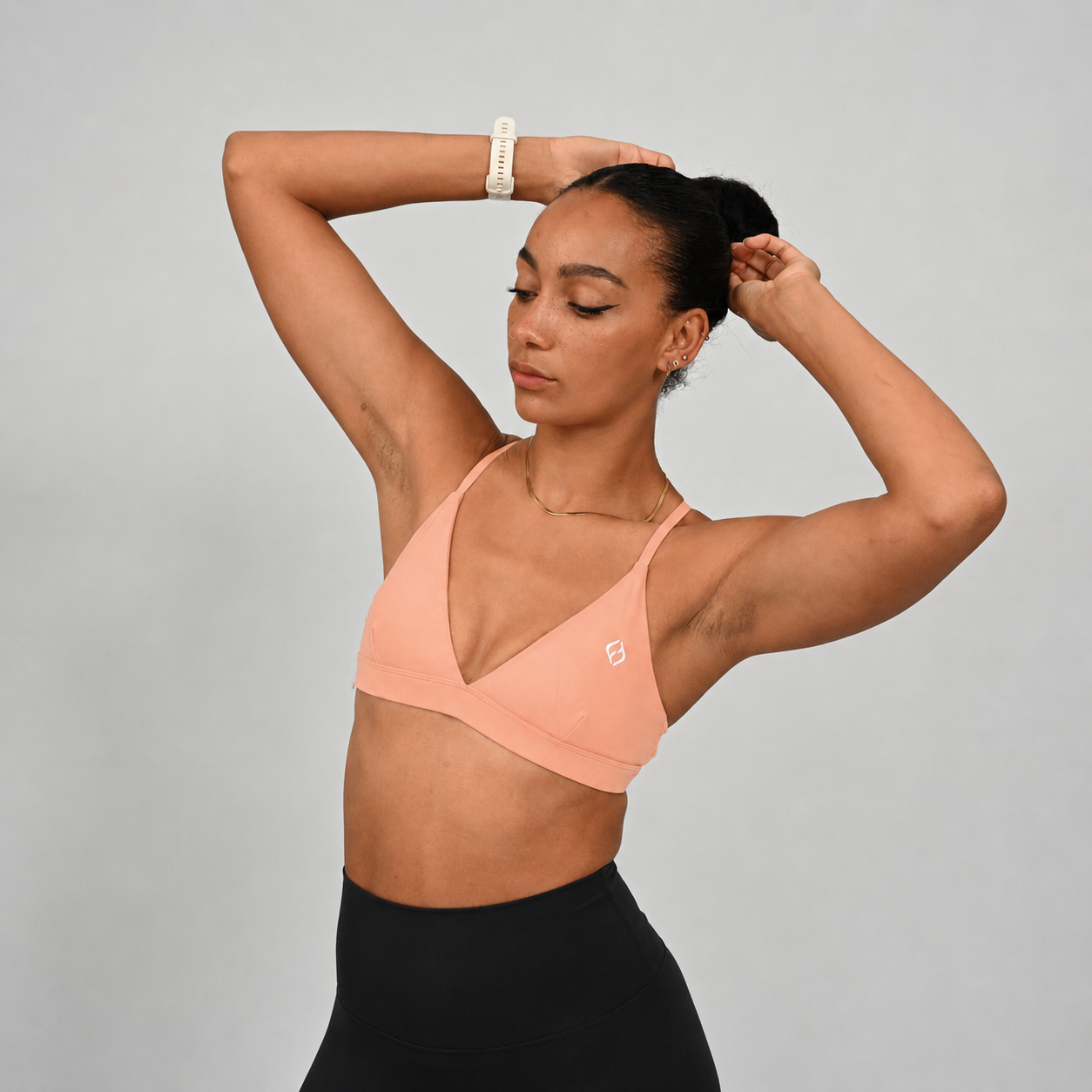A woman wearing a peach-coloured Hybryd crossfit sports bra and black leggings poses with her arms raised against a solid gray studio background.