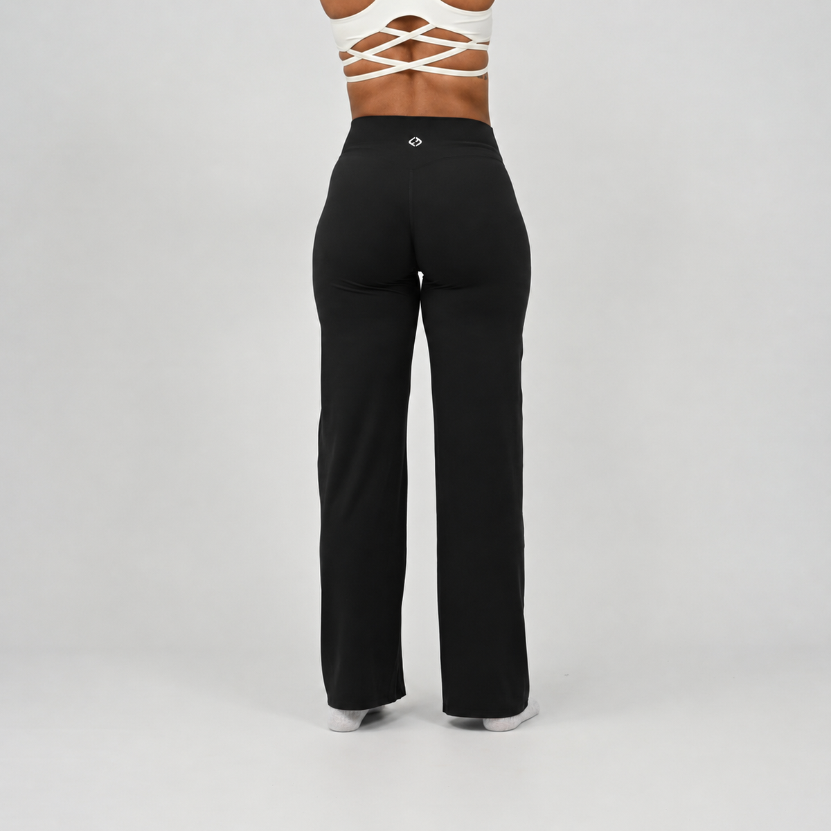 A pilates model wearing black wide-leg pants and white socks, shown from the waist down from the rear against a light gray studio background. From activewear brand Hybryd