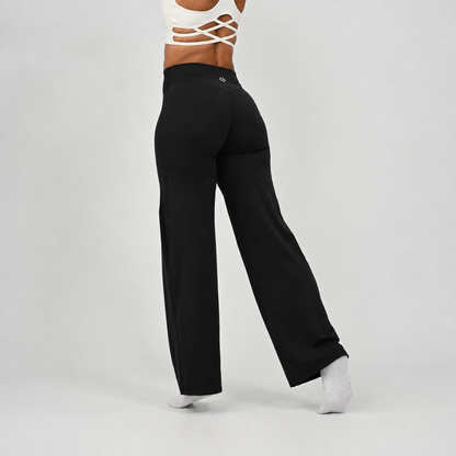 A pilates model wearing black wide-leg pants and white socks, shown from the waist down in a side profile against a light gray studio background. From activewear rand Hybryd