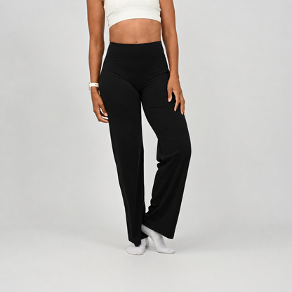 A pilates model wearing black wide-leg pants and white socks, shown from the waist down, from the front against a light gray studio background. From activewear brand Hybryd
