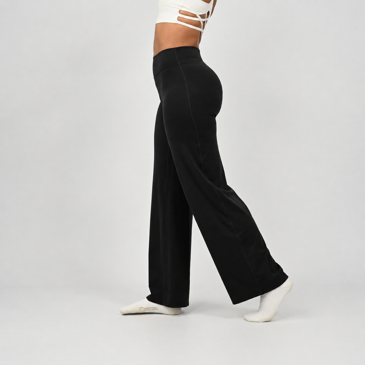 A pilates model wearing black wide-leg pants and white socks, shown from the waist down in a side profile against a light gray studio background. From activewear brand Hybryd