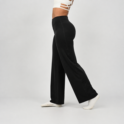 A pilates model wearing black wide-leg pants and white socks, shown from the waist down in a side profile against a light gray studio background. From activewear brand Hybryd