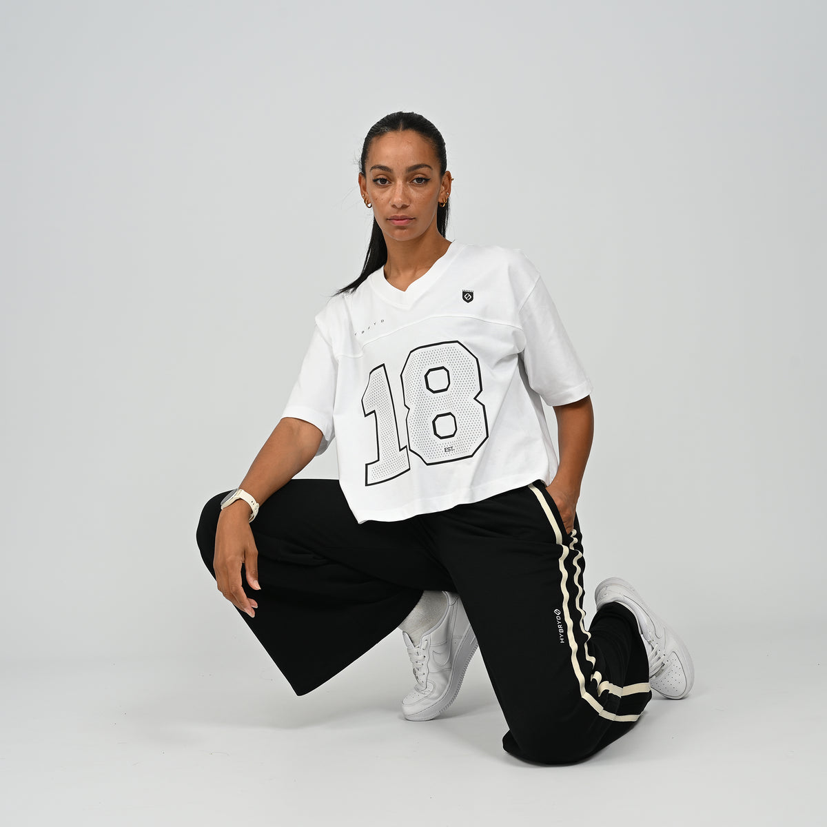 Woman wearing a white oversized collegiate style American football jersey t-shirt by Hybryd paired with dark pants and sneakers, crouching pose