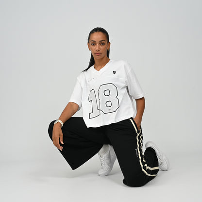 Woman wearing a white oversized collegiate style American football jersey t-shirt by Hybryd paired with dark pants and sneakers, crouching pose
